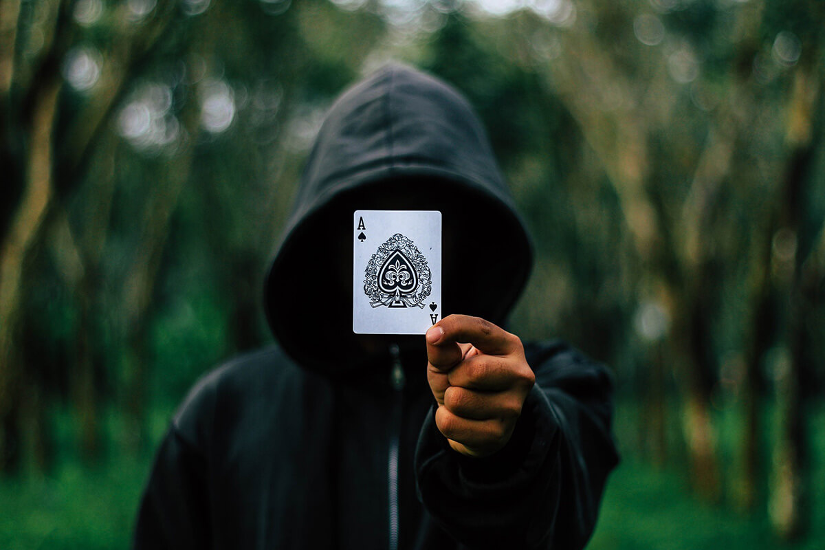 Symbolbild für anonyme Hinweise zu Korruption oder Wirtschaftskriminalität: Person mit scharzem Kapuzenpullover hält Pik-Ass vor Gesicht, Fotocredit iStock.com / AndreyPopov Person mit scharzem Kapuzenpullover hält Pik-Ass vor Gesicht